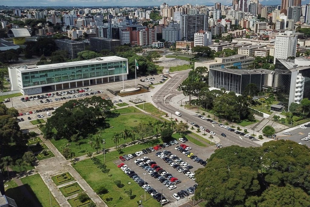 Vista aérea de uma paisagem urbana com uma mistura de edifícios modernos e tradicionais. Estruturas proeminentes incluem dois grandes edifícios flanqueando uma praça aberta com espaços verdes e estacionamentos. Vários carros estão estacionados e edifícios altos erguem-se ao fundo, sugerindo a agitação em torno da sede do TCE – PR.